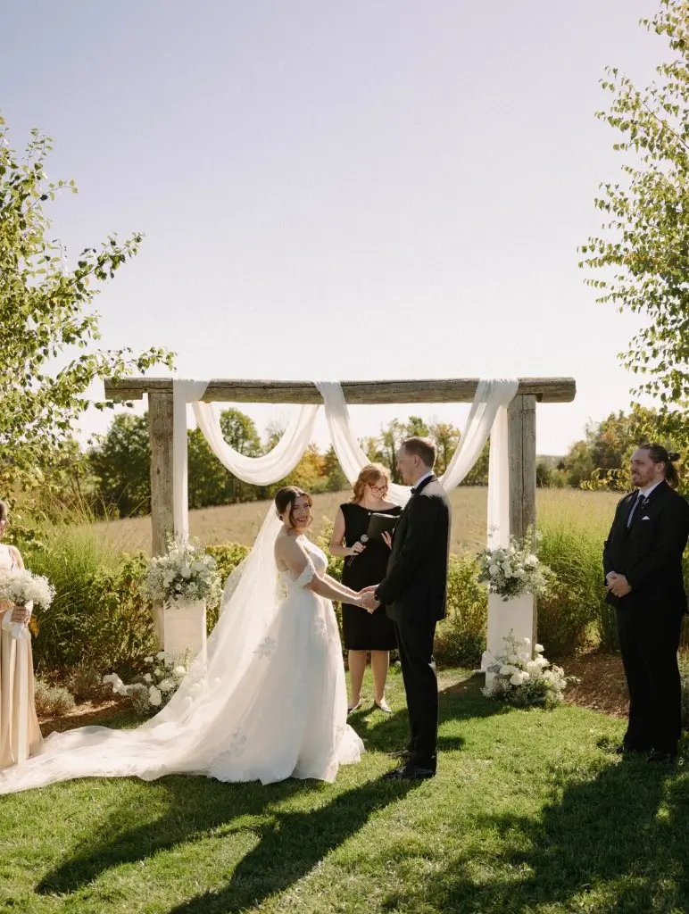 Maureen officiating an outdoor countryside wedding ceremony under a wooden arbour with white draping
